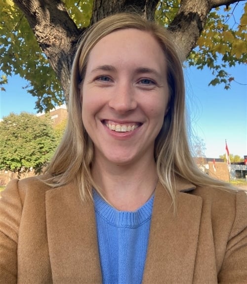 Blond woman in brown blazer and blue shirt smiling with tree in the background