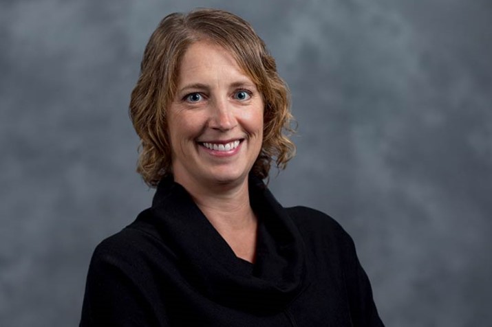 A woman with short, wavy light brown hair wearing a black top smiles at the camera in front of a gray, textured background.