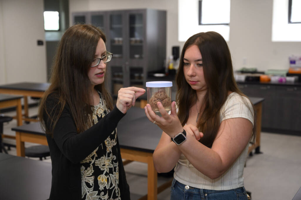 Two women in a classroom examine a brain specimen in a jar. One woman points at the jar while the other holds it, both focused and engaged in discussion. Laboratory tables and cabinets are visible in the background.