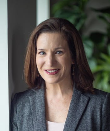 A woman with shoulder-length brown hair smiles while standing indoors. She is wearing a gray blazer over a light top. The background shows green foliage and a white pillar.