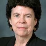 Portrait of a woman with short curly hair wearing a black blazer over a coral top. She has gold earrings and a neutral expression, set against a plain background.