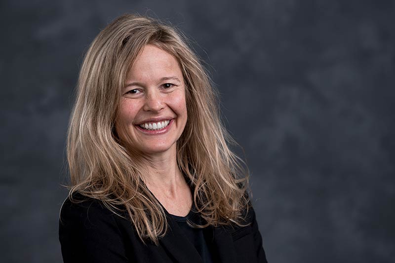 A woman with long, blonde hair is smiling at the camera against a gray, blurred background. She is wearing a black blazer over a dark top.