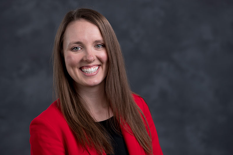 A woman with long brown hair smiling, wearing a red blazer and black top against a blurred dark grey background.