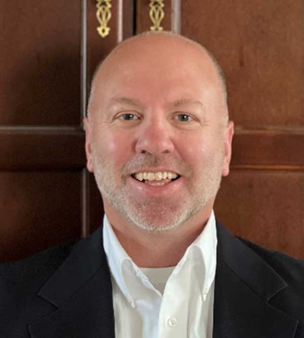 A middle-aged man with a bald head and short white beard, wearing a white dress shirt and a dark suit jacket, smiles warmly at the camera. The background features a wooden paneled wall.