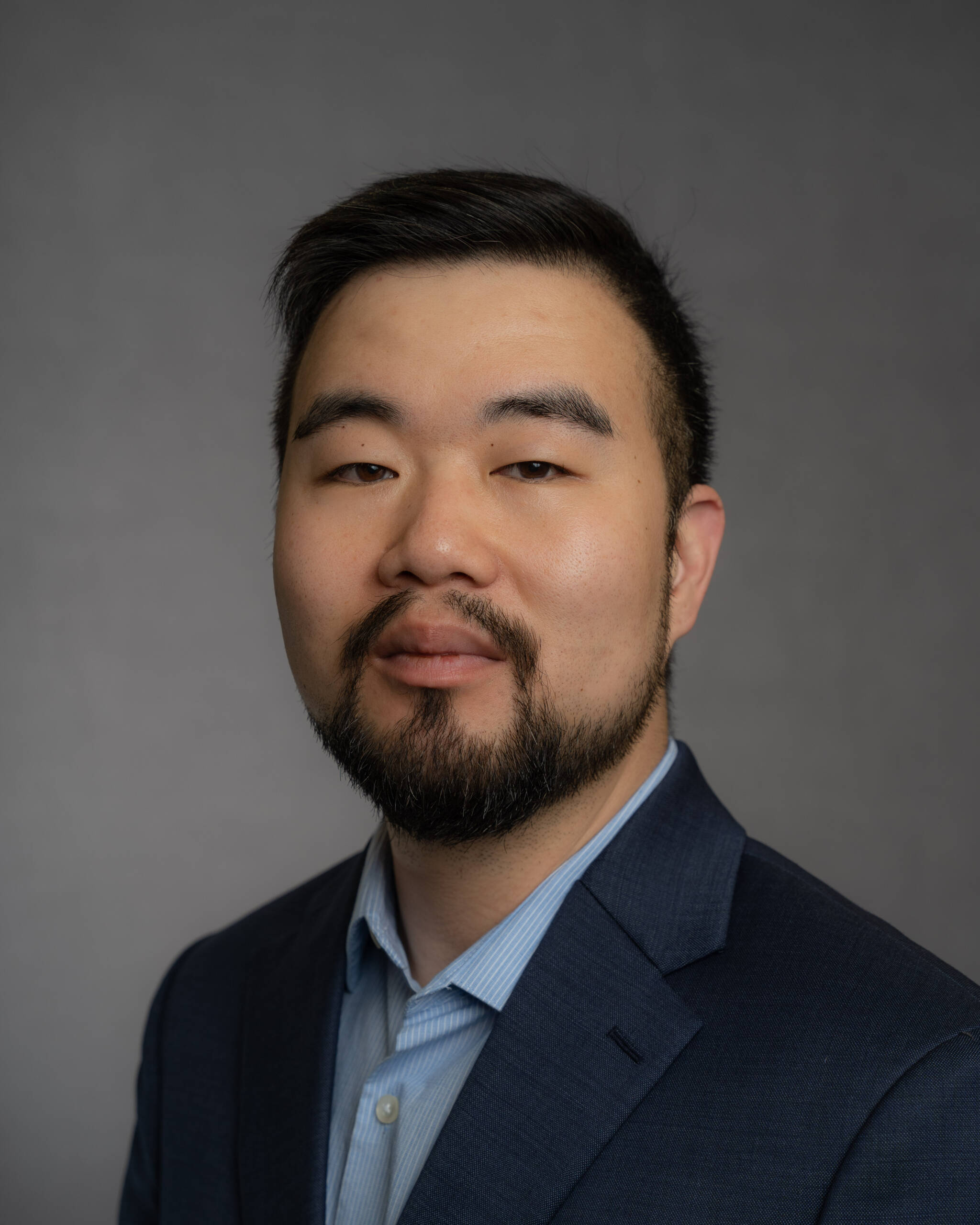A man with short black hair, a beard, and a mustache, wearing a blue suit jacket over a light blue collared shirt, is posing against a plain gray background. He is looking directly at the camera with a neutral expression.
