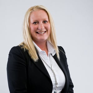 A woman with straight blonde hair smiles, dressed in a black blazer over a white collared shirt. She stands against a plain light-colored background.