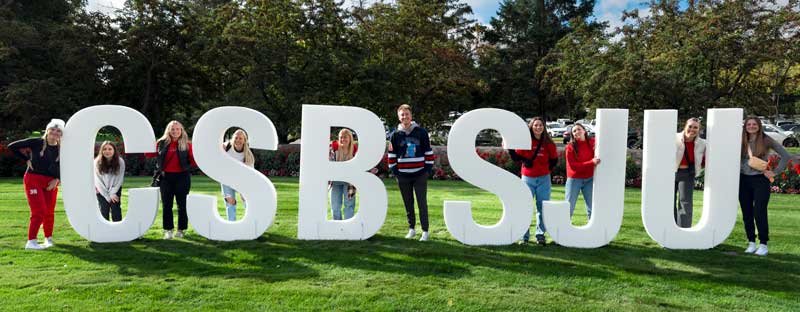 A group of people stand on a grassy lawn, each positioned between large white letters that spell "CSBSJU." Trees and a cloudy sky are visible in the background.