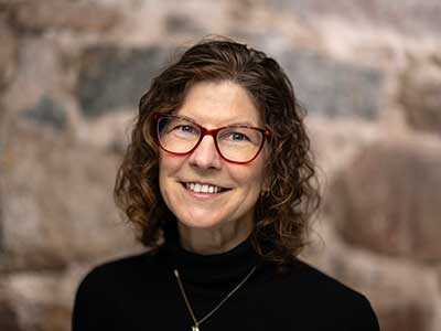 A person with curly hair and red glasses smiles while wearing a black turtleneck. The background is a textured stone wall.