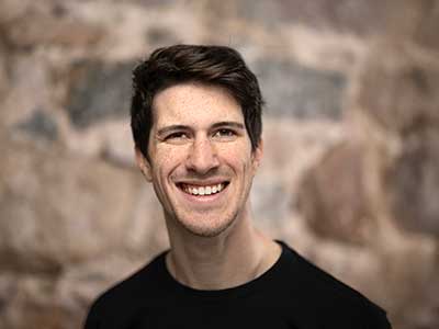 A person with short dark hair and a black shirt is smiling at the camera. The background is a textured stone wall.
