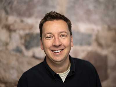 A man with short brown hair, wearing a black shirt, is smiling at the camera. The background is a blurred stone wall.