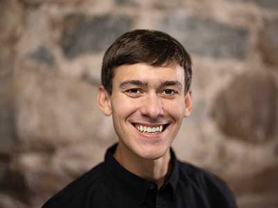 A young man with short dark hair smiling broadly, wearing a black shirt. He is in front of a blurred stone wall background.