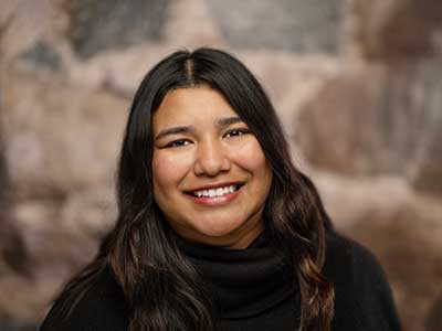 Smiling person with long dark hair wearing a black turtleneck, in front of a blurred neutral background.