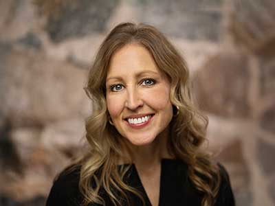 Smiling woman with long, wavy blond hair wearing a black top, standing against a stone wall background.