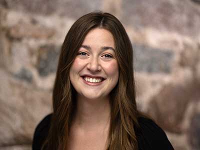 A person with long brown hair is smiling in front of a blurred stone wall background. They are wearing a black top.