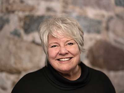 A person with short gray hair smiles warmly while wearing a black top. The background is a stone wall with various shades of gray and brown.