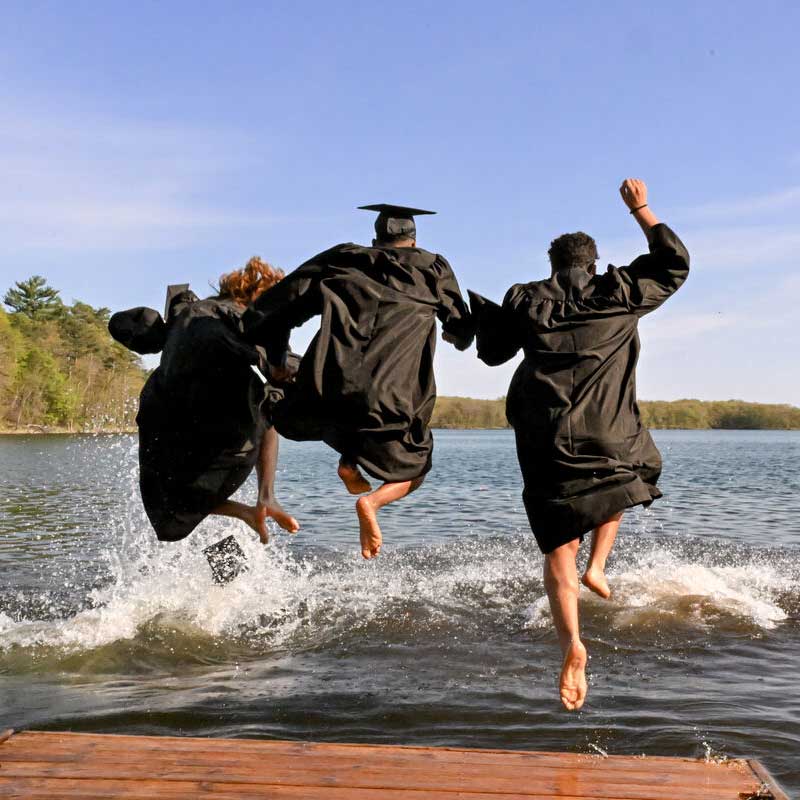 Three graduates in caps and gowns joyfully jump into a lake from a wooden dock, creating splashes in the water, under a clear blue sky with a backdrop of trees.
