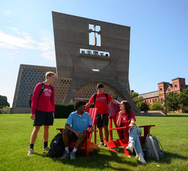 Four students are gathered in conversation, sitting and standing around red chairs on a grassy lawn. The background features a modern building with a geometric design and a brick building under a clear blue sky.