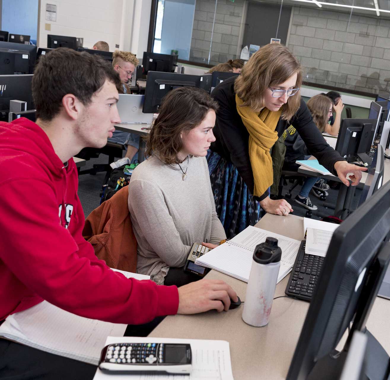 A teacher helps two students in a computer lab. The students are seated at computers, one wearing a red sweatshirt and the other a gray sweater. The teacher points at the screen while the students focus on their work.