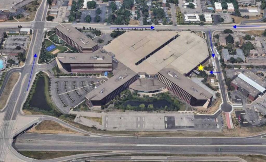 Aerial view of a large, X-shaped building complex with parking lots, surrounded by roads and neighboring structures. There's a yellow star on the right side of the image, indicating a specific location. Trees and smaller buildings are visible nearby.
