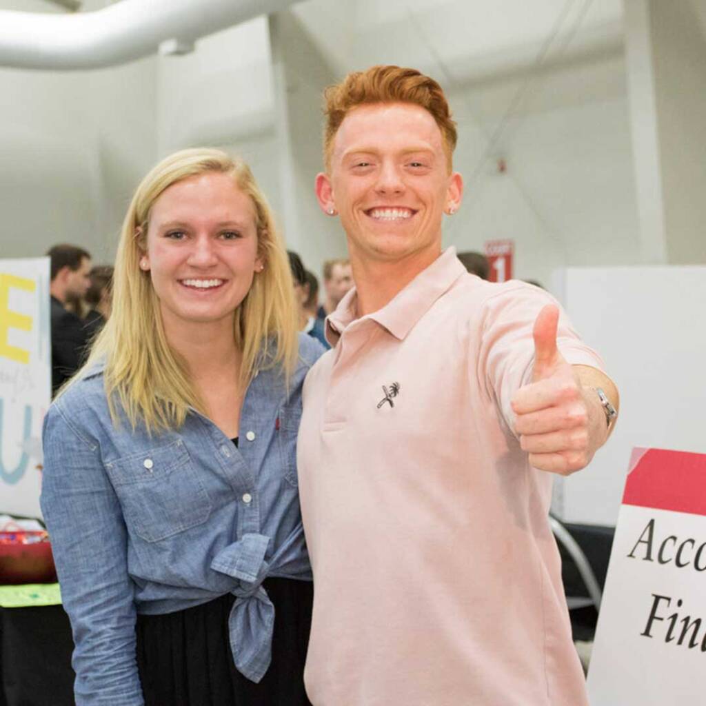 Two people smiling indoors; a woman on the left with long blonde hair wearing a blue shirt, and a man on the right with short red hair in a light pink polo shirt giving a thumbs-up. People and signs are blurred in the background.