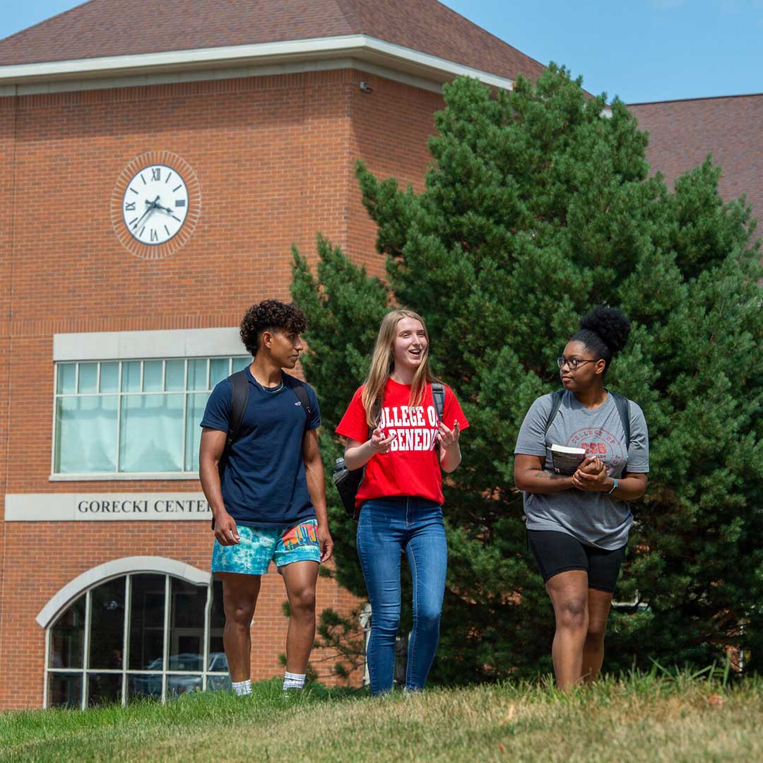 Three students walk and chat outside Gorecki Center, a brick building with a clock and large windows, on a sunny day. Two students carry notebooks, and one wears a red "College of Saint Benedict" shirt.