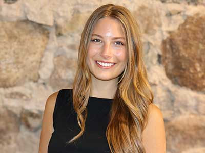 A woman with long, wavy, light brown hair wearing a sleeveless black top smiles in front of a stone wall background.