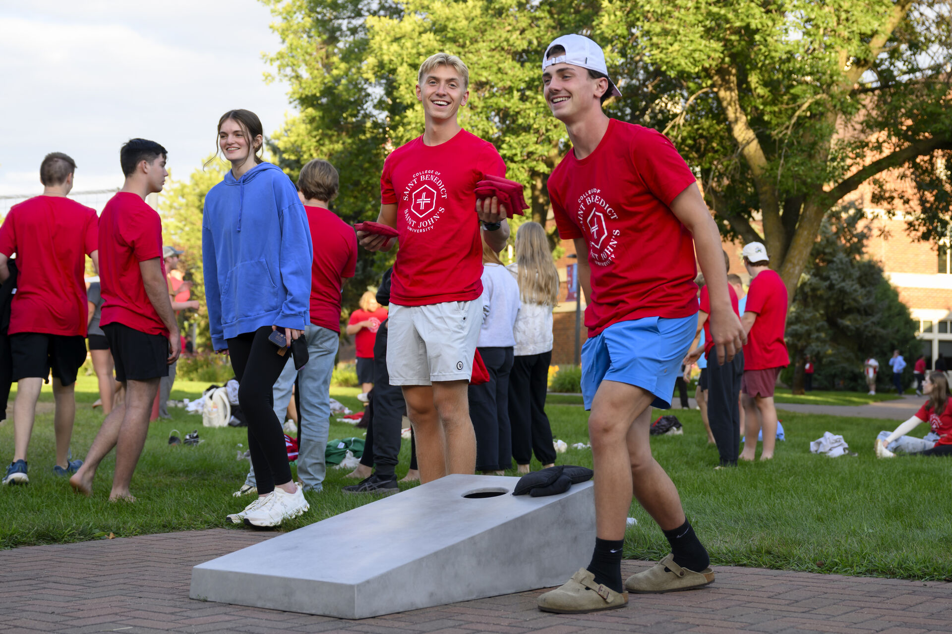 A group of young people play cornhole outside on a sunny day. Two smiling boys in red shirts and shorts toss bean bags, while others watch and chat in the background on the grassy lawn.