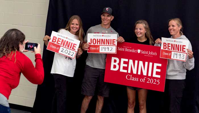 Four people pose for a photo, each holding signs showing their graduation years from Saint Benedict and Saint John’s, including classes of 1992, 1993, 2022, and 2025. A fifth person is taking their picture.