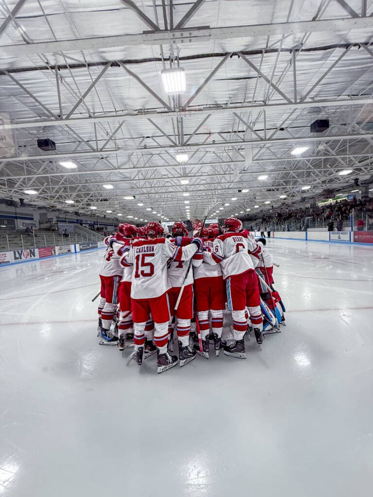 A hockey team in red and white uniforms huddles together on the ice rink before a game, with empty stands on one side and spectators on the other, under bright arena lights.