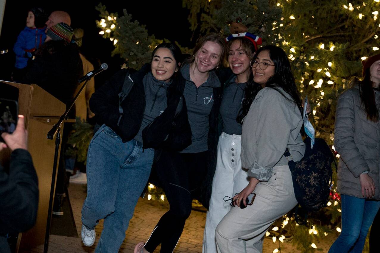 A group of people poses happily for a photo in front of a microphone stand and a large, decorated tree with lights. Others are seen in the background, some taking photos, all wearing casual winter outfits.