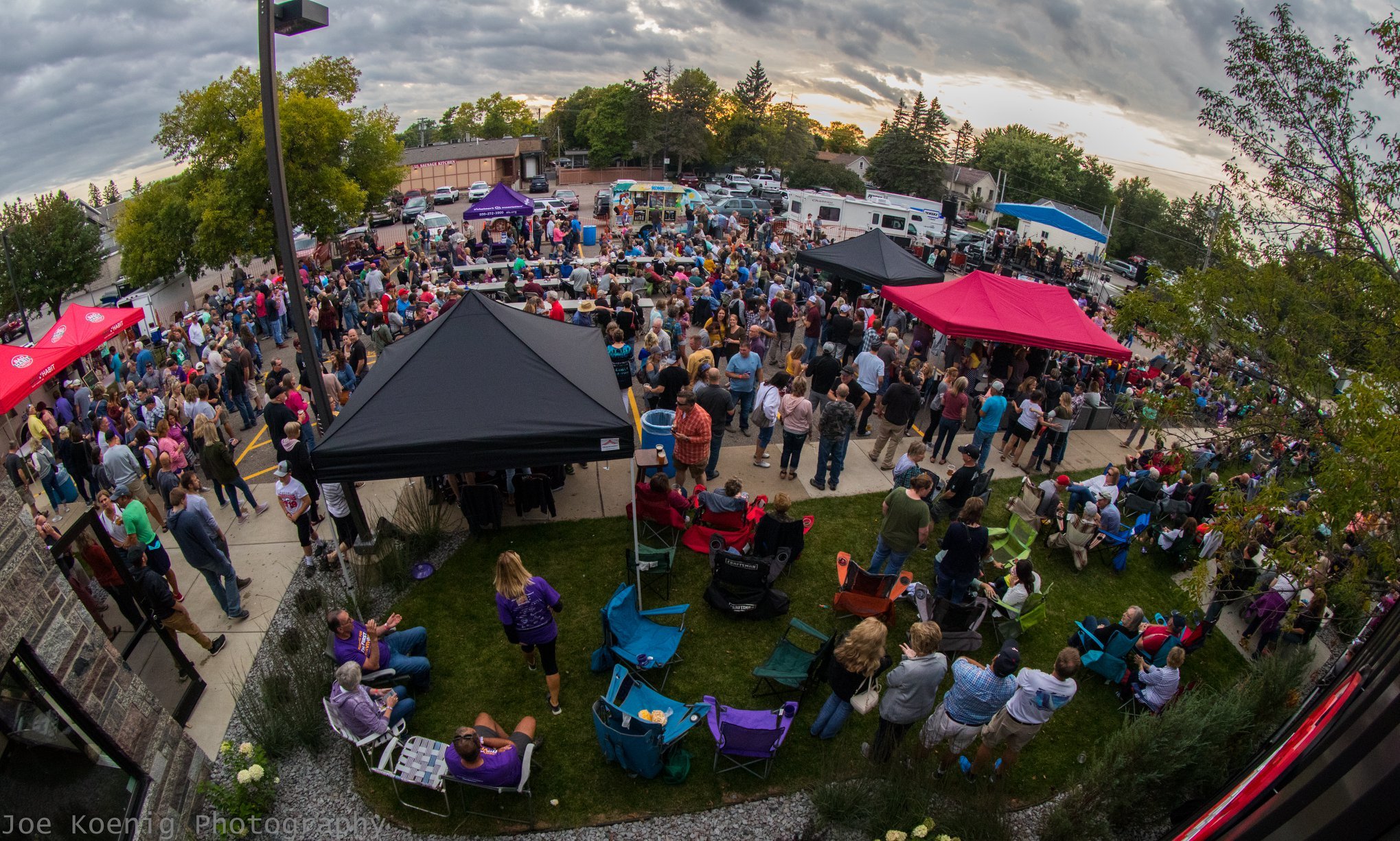 A large outdoor gathering with people standing and sitting under various colored canopies with tables. It's a lively scene, suggesting a community event or party, surrounded by trees and a few buildings. Cloudy sky overhead.