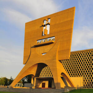 A modern church with bold geometric architecture, featuring a large honeycomb-patterned wall and a tall rectangular bell tower with a cut-out cross and arched base, set against a clear sky.