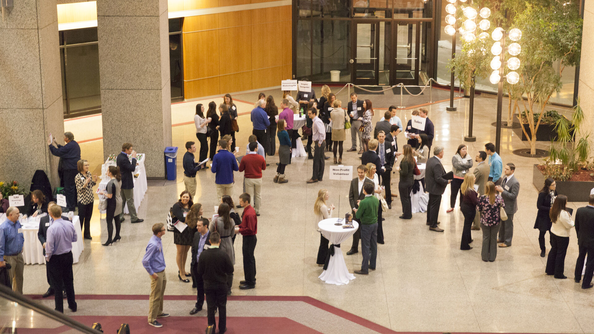 Aerial view of a networking event in a spacious lobby. Groups of people are gathered around high-top tables, engaging in conversations. Some tables have signs on them. The space is well-lit with modern decor and tall plants.