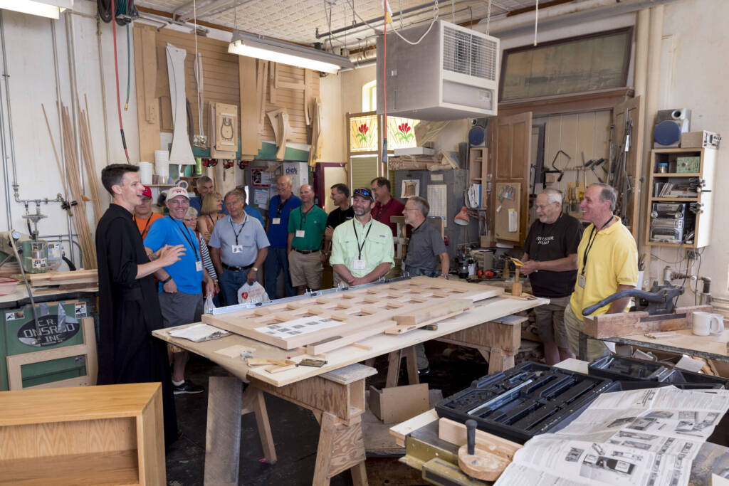 A group of people stand and listen to a man speaking in a woodworking shop, surrounded by tools, unfinished wood projects, and workbenches under bright indoor lighting.