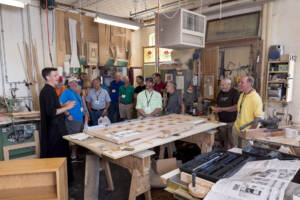 A group of people stand and listen to a man speaking in a woodworking shop, surrounded by tools, unfinished wood projects, and workbenches under bright indoor lighting.