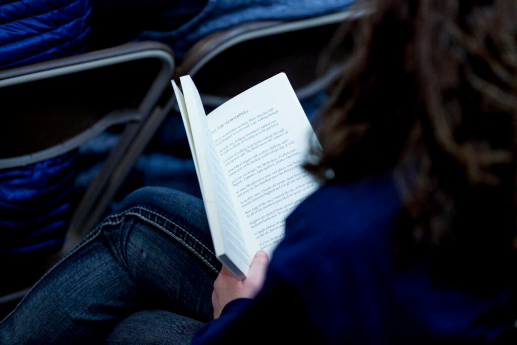 A person with long hair, dressed in dark clothing, is sitting and reading a book. The photo is taken from behind, focusing on the open pages of the book and the reader’s hand. Rows of chairs are visible in the background.