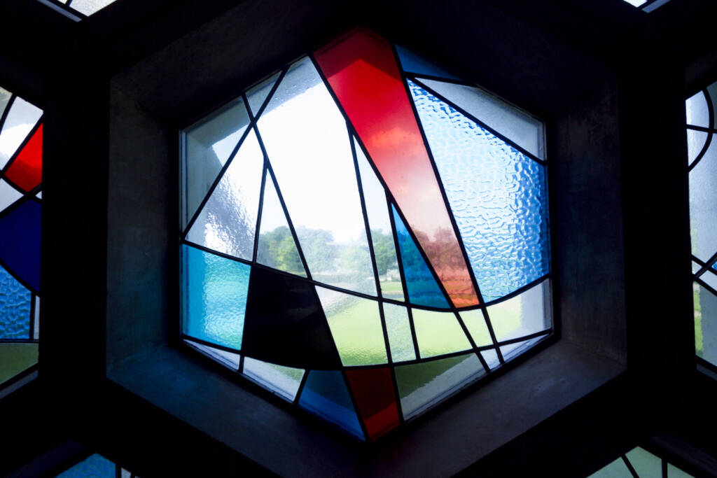 Hexagonal stained glass window with abstract geometric shapes in red, blue, white, and black. Light shines through, illuminating the colors and casting a view of greenery outside in the background.