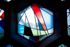 Hexagonal stained glass window with abstract geometric shapes in red, blue, white, and black. Light shines through, illuminating the colors and casting a view of greenery outside in the background.