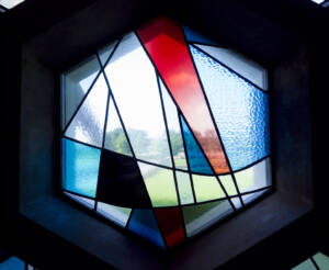 Hexagonal stained glass window with abstract geometric shapes in red, blue, white, and black. Light shines through, illuminating the colors and casting a view of greenery outside in the background.