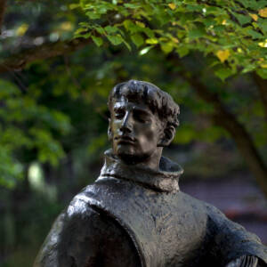 Bronze statue of a man in a monk’s robe holding an open book, with a stack of books at his side, standing outdoors among trees and fallen leaves.