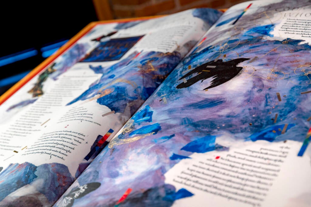 An open illustrated book with vibrant blue, purple, and black abstract artwork and stylized text on both pages, resting on a flat surface with a blurred brick background.