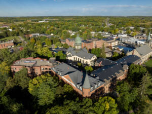 Aerial view of a large, historic brick building complex with a green dome, surrounded by trees and a small town under a clear blue sky.