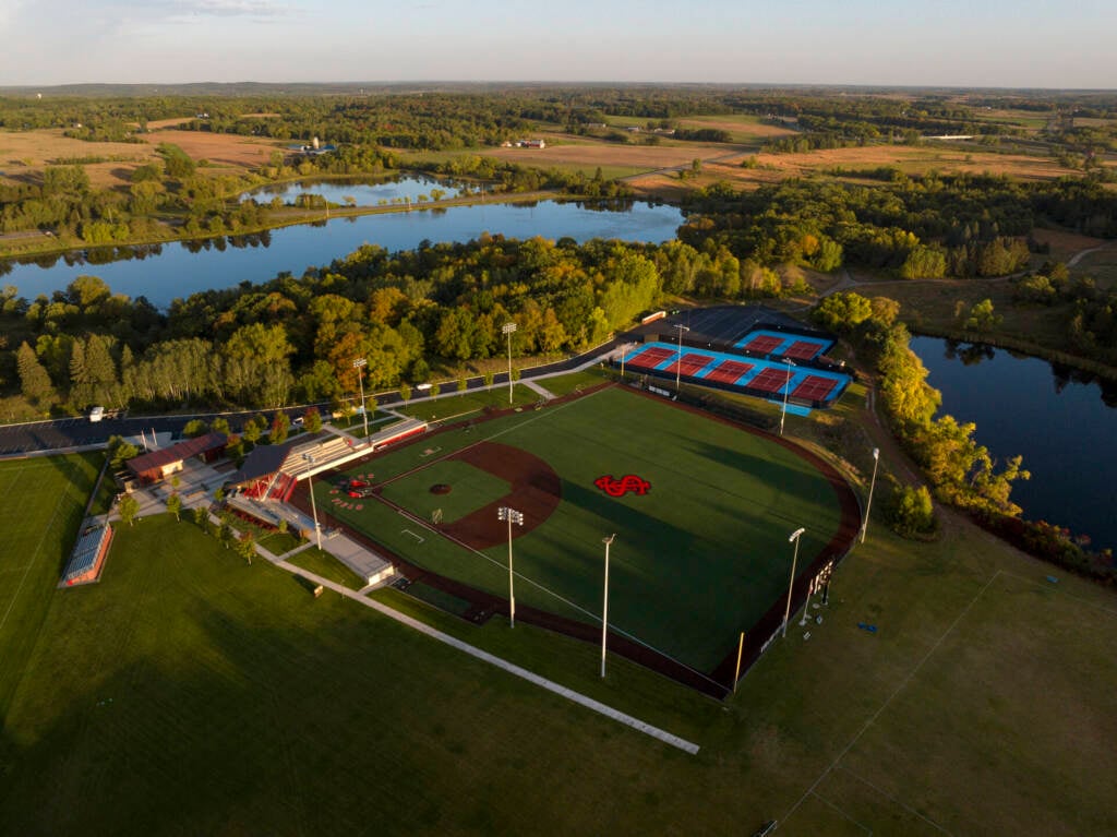 Aerial view of a baseball field and adjacent tennis courts surrounded by trees and ponds, with farmland and a river in the background under a clear sky.