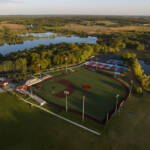 Aerial view of a baseball field and adjacent tennis courts surrounded by trees and ponds, with farmland and a river in the background under a clear sky.