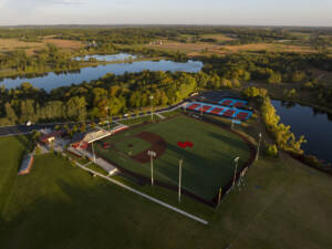 Aerial view of a baseball field and adjacent tennis courts surrounded by trees and ponds, with farmland and a river in the background under a clear sky.