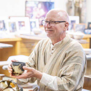 An older man with glasses and a striped beige shirt sits at a wooden table, smiling and holding a ceramic mug. Art and pottery pieces are displayed in the background.