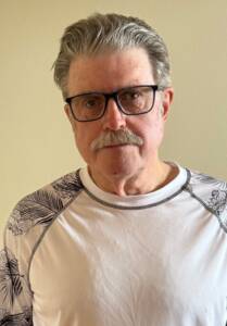 An older man with gray hair, a mustache, and glasses is wearing a white shirt with black patterned sleeves. He stands in front of a plain beige wall, looking directly at the camera with a neutral expression.