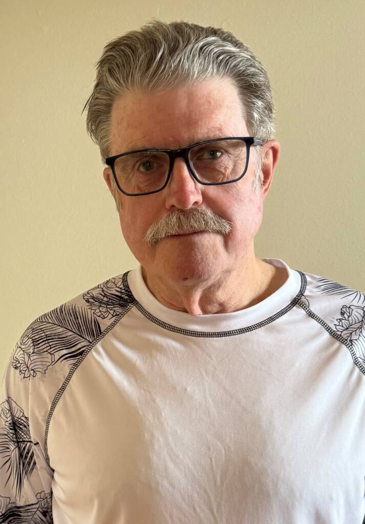 An older man with gray hair, a mustache, and glasses is wearing a white shirt with black patterned sleeves. He stands in front of a plain beige wall, looking directly at the camera with a neutral expression.