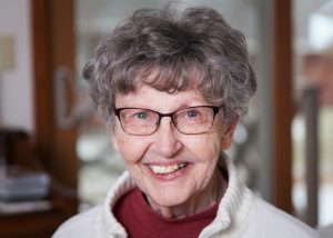 An elderly woman with short, curly gray hair and glasses smiles warmly at the camera. She is wearing a white sweater and a red turtleneck. The background is softly blurred, suggesting an indoor setting.