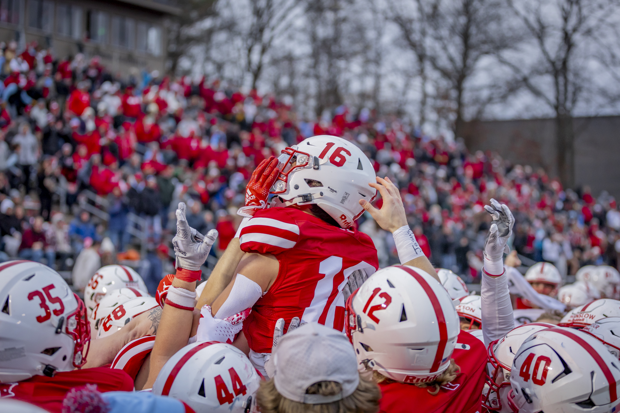 A group of football players in red and white uniforms lift a teammate wearing number 16 into the air as a crowd of fans watches in the background on a cloudy day.